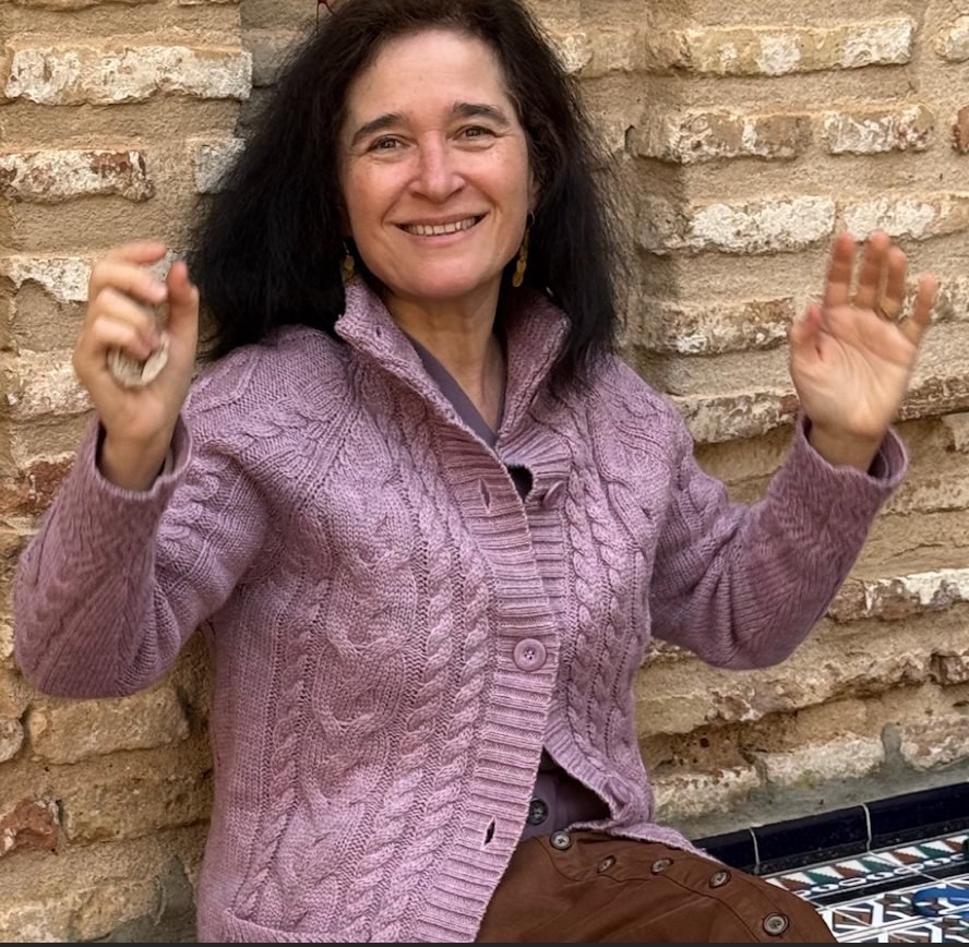 Color portrait of Ági Vető — a woman with shoulder-length curling dark hair and gold drop earrings, smiling warmly at the camera with both hands raised palms-out at chest height in a welcoming gesture, wearing a heathered dusty-pink cable-knit cardigan over a mauve shirt, seated against a weathered exposed-brick wall with a strip of tilework visible at the lower edge.