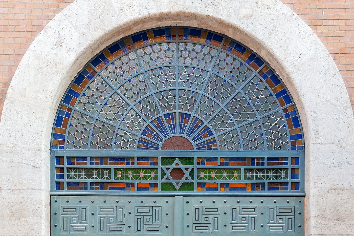Photograph of the iron-and-stained-glass arched main door of the Kazinczy Street Synagogue in Budapest: a tall semicircular fanlight glazed in a sunburst pattern of cobalt, ochre, white and green tiles set in a slate-grey leaded armature, framed by a smooth limestone arch against red brick; the doors below are heavy panelled metal with geometric maze-like reliefs and a small Star of David at the lintel.