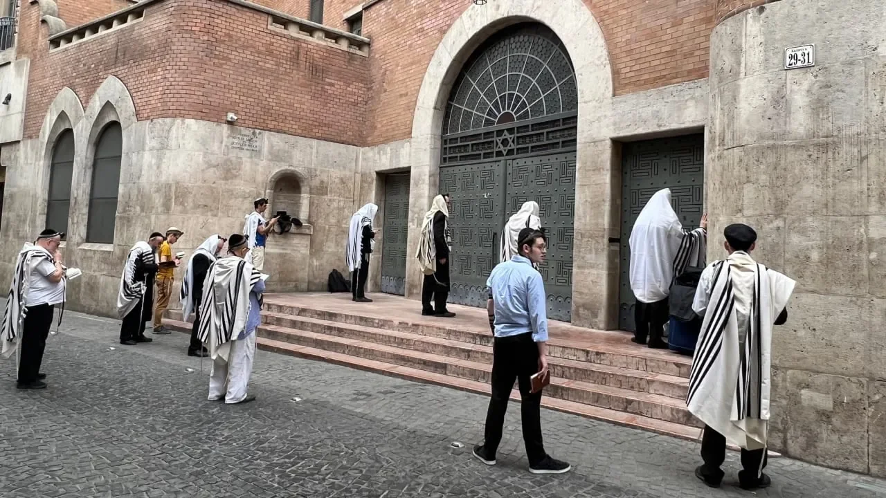 Color photograph of the same Kazinczy Street Synagogue exterior taken from across the cobblestone street: a dozen men in tallitot — most wrapped over the head — stand and pray on the stone steps and sidewalk in front of the closed doors, two leaning into the wall, others holding siddurim, one young man in a white shirt looking back over his shoulder toward the camera.
