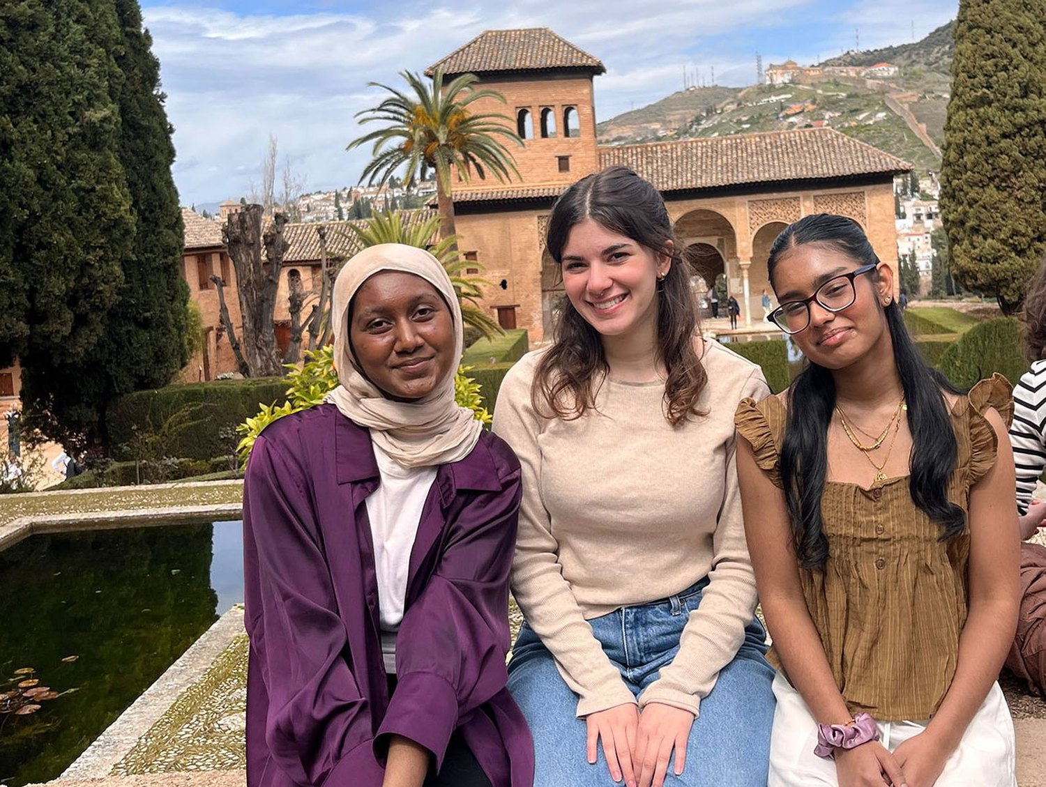 Three Vassar students sit shoulder-to-shoulder on a low marble parapet in front of a long reflecting pool, with the brick-and-tile Alhambra Partal pavilion and arches behind them, a tall palm rising on the left and the Sierra Nevada in the distance: a young woman in a cream hijab and aubergine open coat, a young woman in a beige long-sleeved sweater and jeans with shoulder-length brown hair, and a young woman in a mustard-yellow ruffled top with long dark hair and glasses, all smiling at the camera.